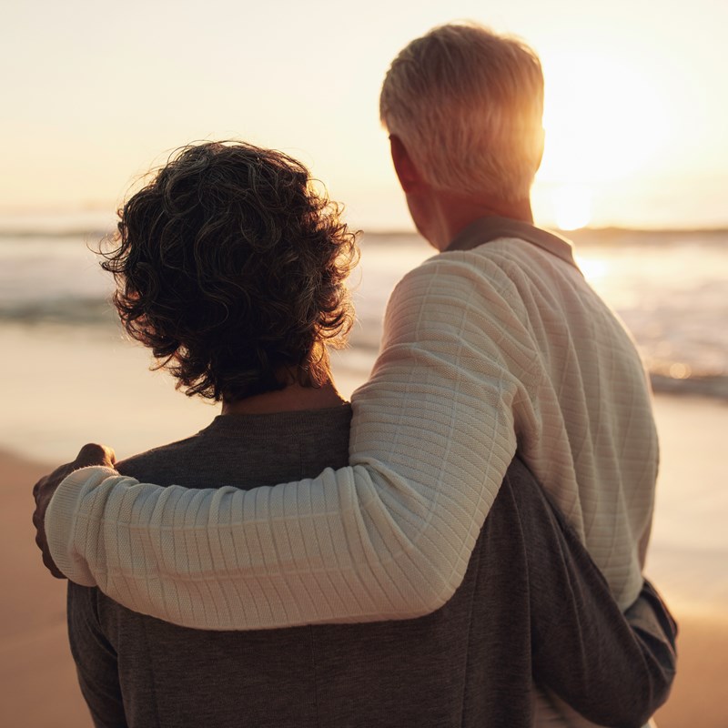 Man och kvinna som står på en strand med ryggen mot fotografen och tittar på solnedgången